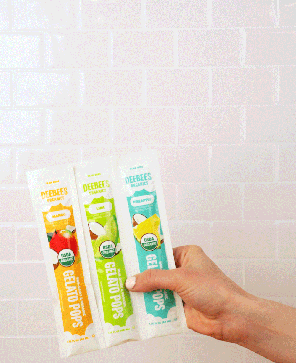 A hand shaking DeeBees Organics Non-Dairy Gelato Pops against a white tile background