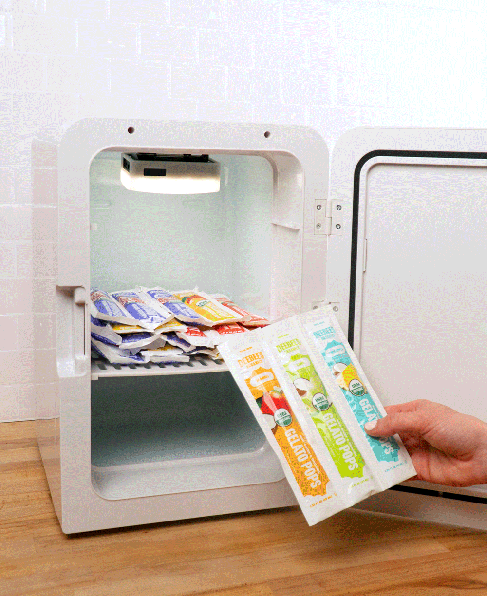 A hand placing DeeBees Organics Non-Dairy Gelato Pops into a small white freezer on a wooden counter against a white tile background