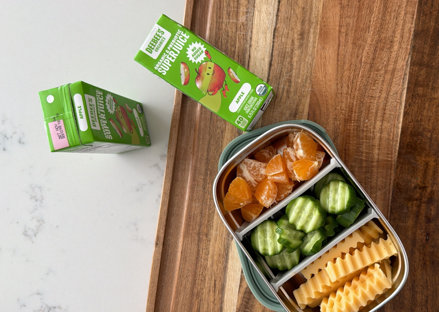 DeeBee's Organics Apple SuperJuice Prebiotic Juice Boxes beside a balanced snack container filled with cucumber slices, orange segments, and crinkle-cut cheese sticks on a wooden board.