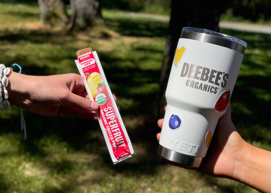 Hand holding a DeeBee’s Organics SuperFruit Strawberry Lemon freeze pop next to a white Yeti branded tumbler with fruit stickers outdoors.