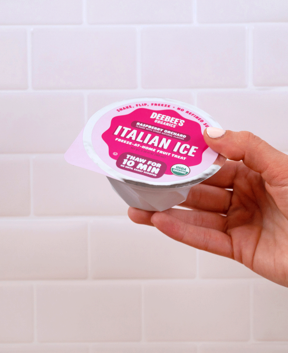 A hand shakes a DeeBee's Organics Italian Ice cup against a white tile background.
