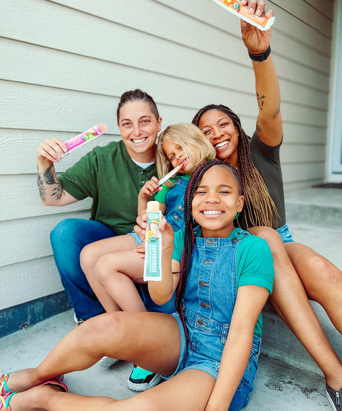 A family comprised of two moms and their two children are sitting on their front steps, smiling, and holding DeeBees Organics SuperFruit Freezies.